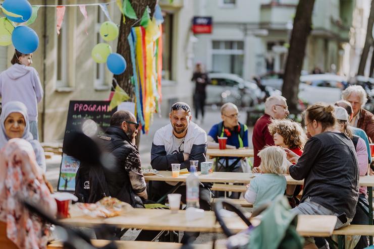 Menschen sitzen an Holztischen, genießen Essen und Getränke bei einer Veranstaltung im Freien mit bunten Dekorationen.